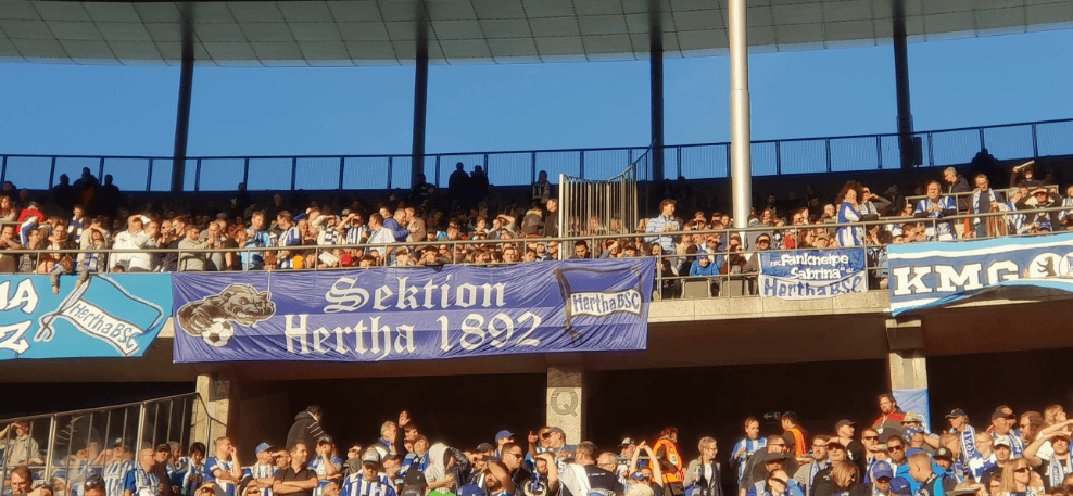 Menschenmenge im Stadion mit Hertha-Banner, blau-weiße Kleidung, sonniges Wetter.