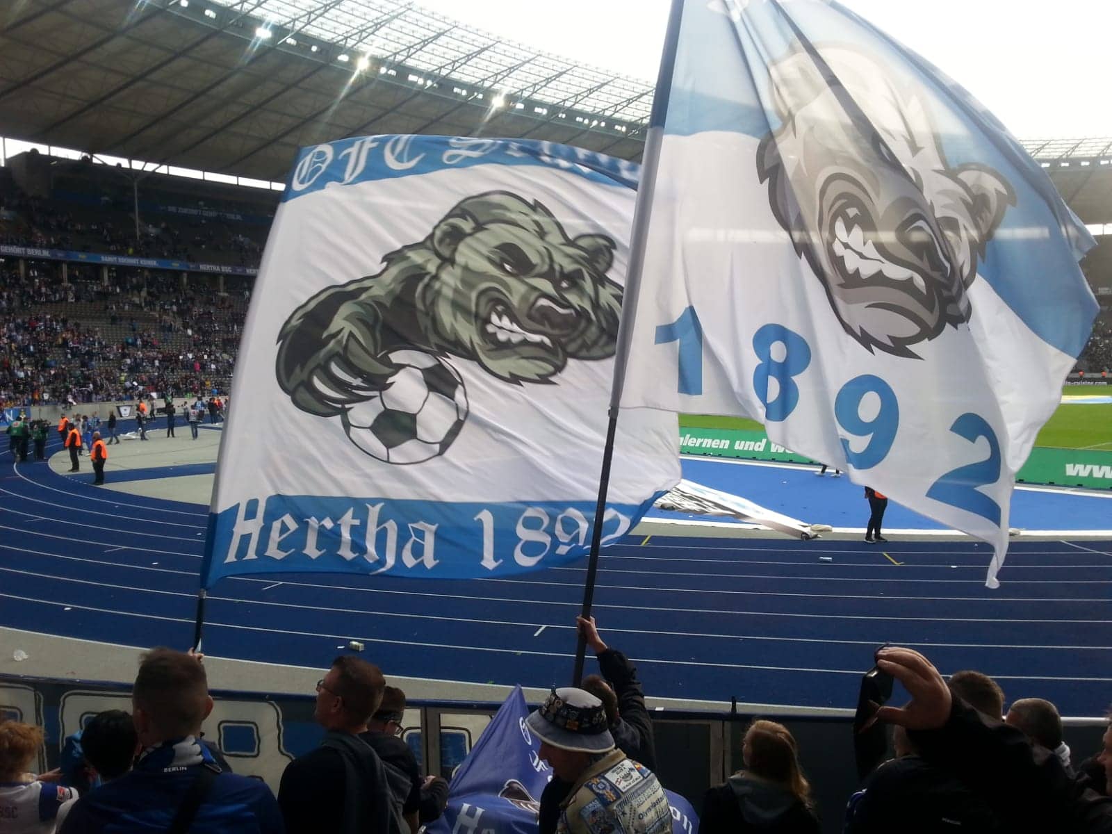 Zwei Fans schwenken Hertha BSC-Fahnen im Olympiastadion; blaue Laufbahn und begeisterte Zuschauer.