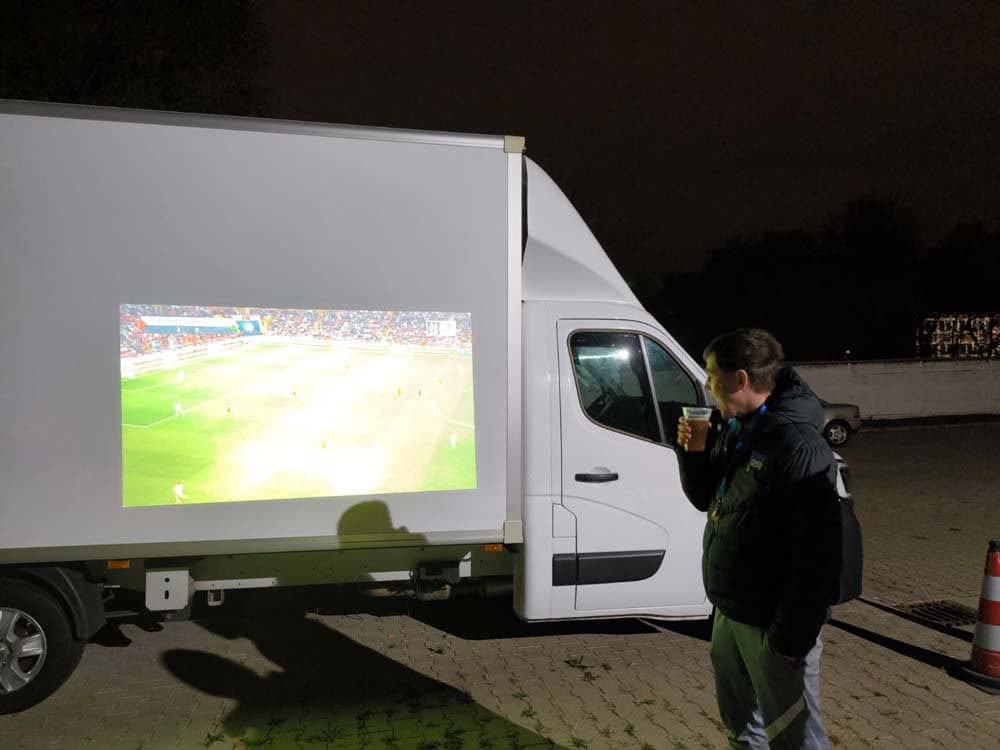 Ein Mann schaut ein Fußballspiel auf einem Lieferwagen, dunkle Kleidung, draußen bei Nacht.