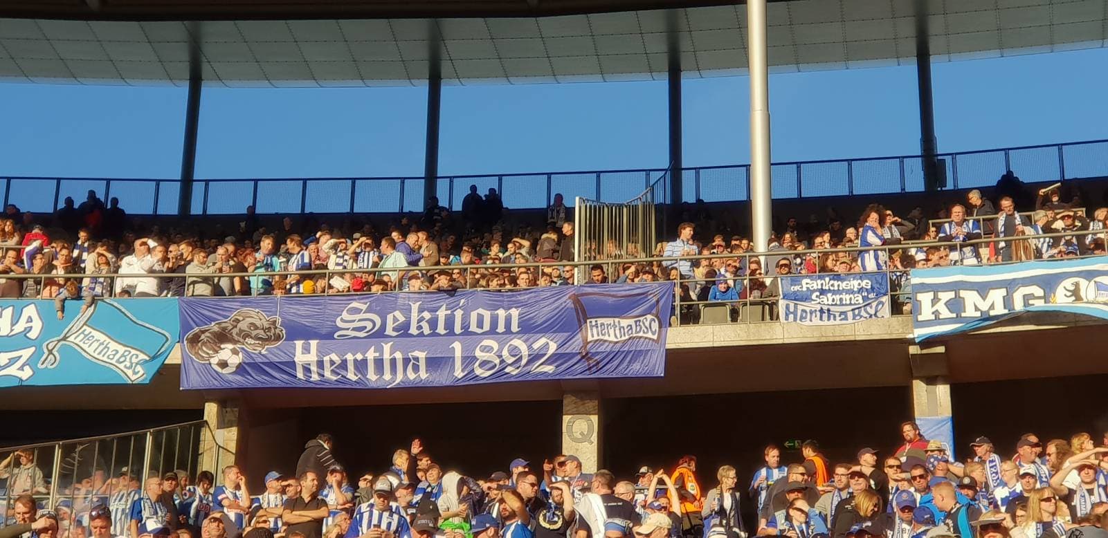 Zuschauer im Stadion tragen blau-weiße Kleidung, fröhlich, unter Bannern wie "Sektion Hertha 1892".