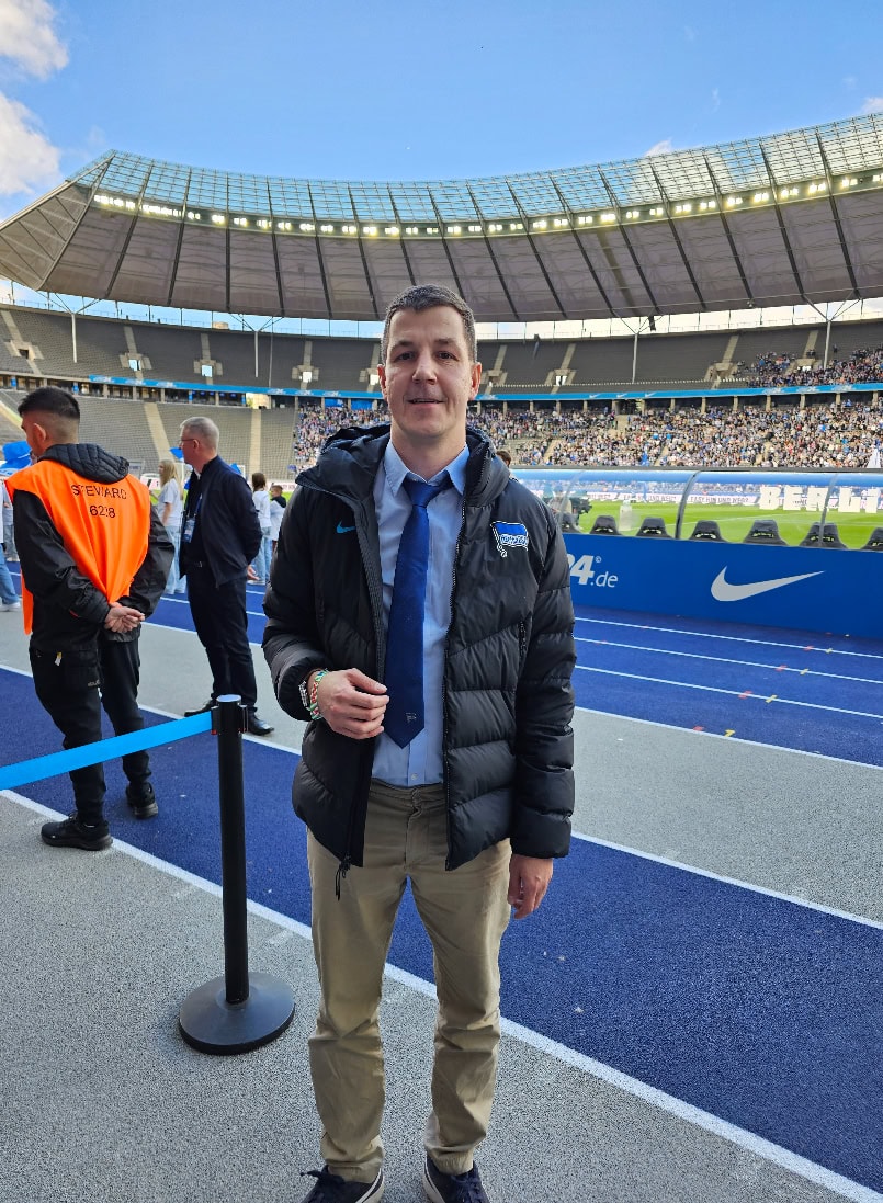 Ein Mann lächelt im Stadion, trägt eine schwarze Jacke und Khakihose. Im Hintergrund Sicherheitskräfte.